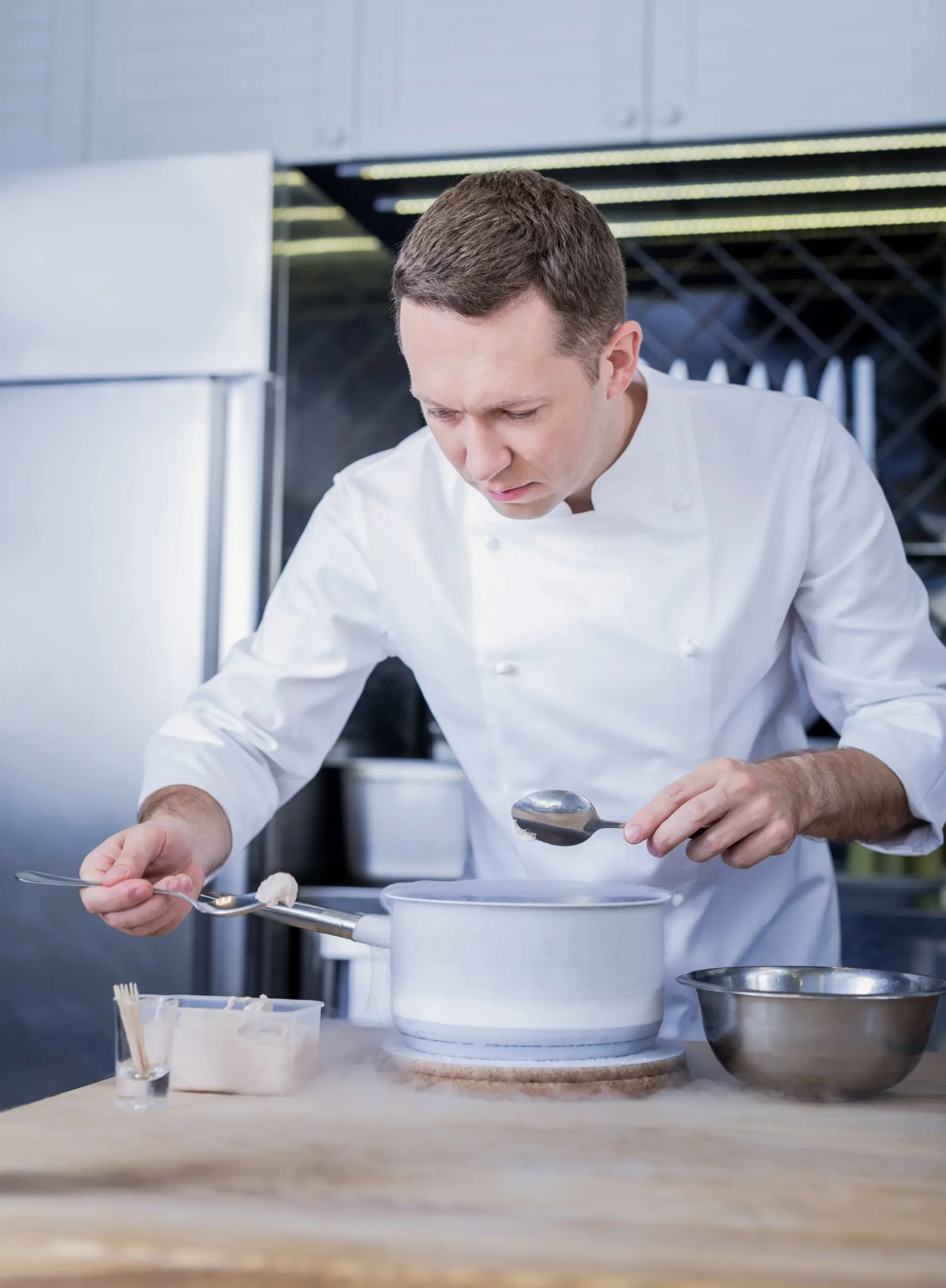 A professional private estate chef in a crisp white uniform and apron basting a whole roasted fish in a high-end, minimalist kitchen equipped with a professional Gaggenau vent hood