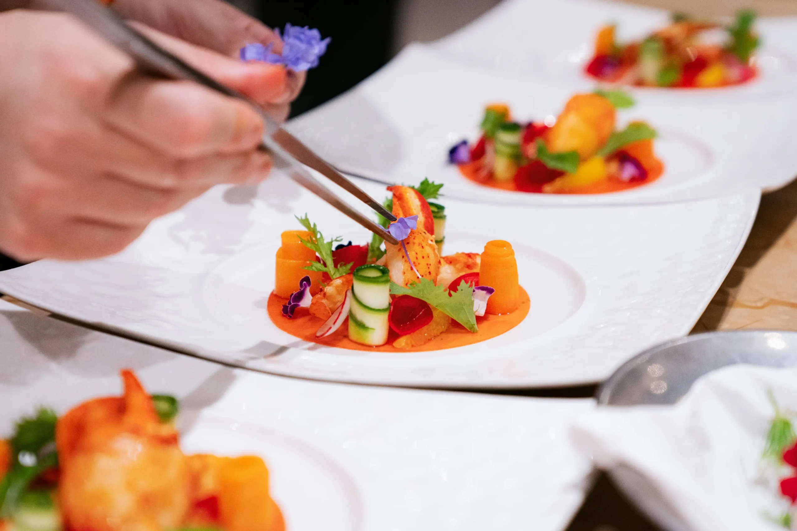 A close-up of a Michelin-caliber chef using precision tweezers to plate an intricate lobster dish with edible flowers and fresh vegetables, showcasing technical culinary artistry